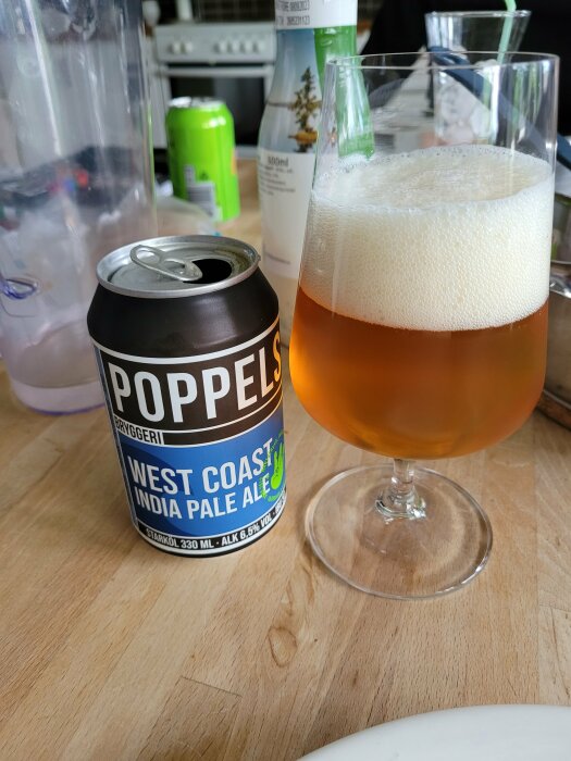 Poppels West Coast IPA burk, ölglas med skum, matsalsbord, suddiga föremål i bakgrunden.