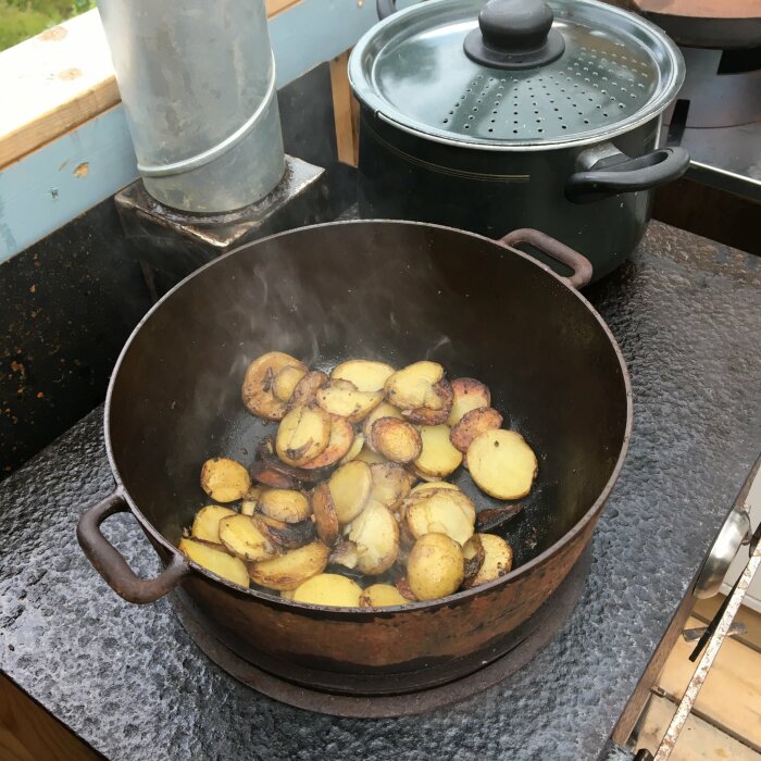 Stekt potatis i gjutjärnspanna på utomhus kök, ångande varm, med kokkärl i bakgrunden.