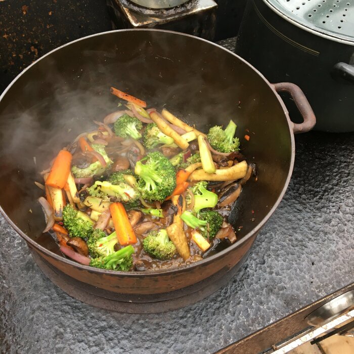 Wokade grönsaker med broccoli och morötter i ångande, brun stekpanna på utomhuskök.