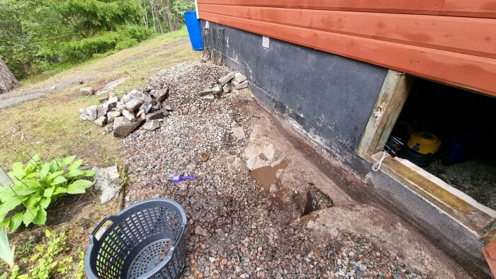 Grund av hus, grus, öppen krypgrund, stenar, blommor, skogsbakgrund. Hink, skovel, trädgårdsarbete eller grundreparation.