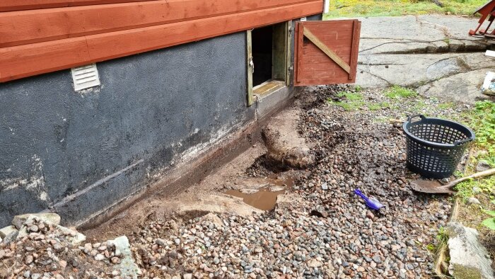 Grävning vid husgrund, öppet kryputrymme, spade, korg, stenar, jord.