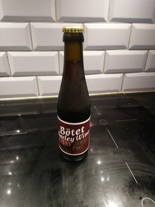 En flaska öl med etiketten "Bötet Barley Wine" står på en svart yta med kakelvägg i bakgrunden.