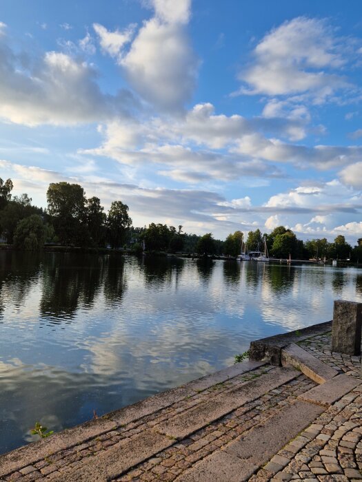 Tranquility at lakeside, cobblestones, trees, clouds reflection in water, blue sky, serene outdoors.