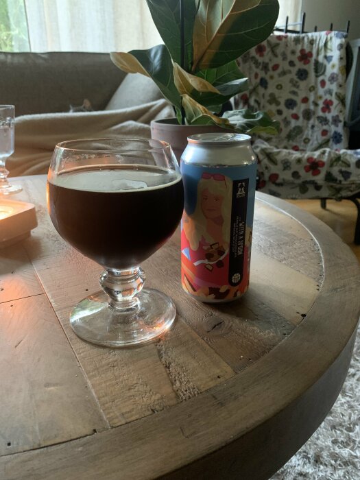 Ett glas mörkt öl och en ölburk på ett träbord, interiör med soffa och växt.