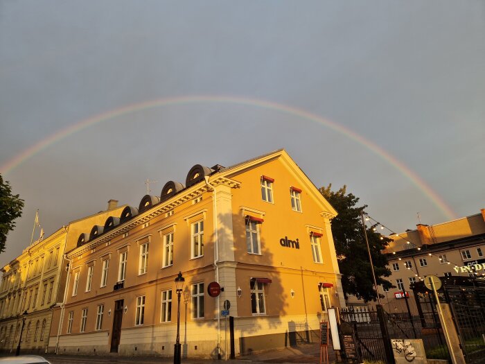 Regnbåge över byggnader i solnedgång med våta gator, vilket antyder nylig regn.