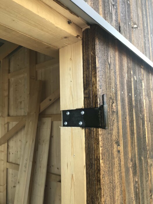 En hörnbeslag fäster en träbalk till en vägg av väderbitat trä under en taköverhäng.