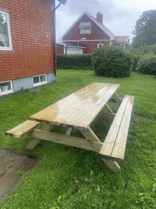 En tom träpicknickbord på en gräsmatta framför en röd tegelvägg, ser regnigt och molnigt ut.