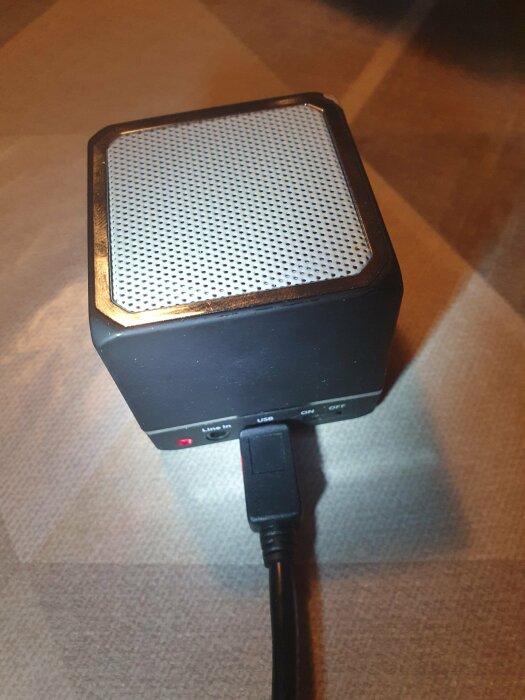 Liten svart högtalare ansluten med USB-kabel, röd indikatorlampa tänd, står på brunt underlag.