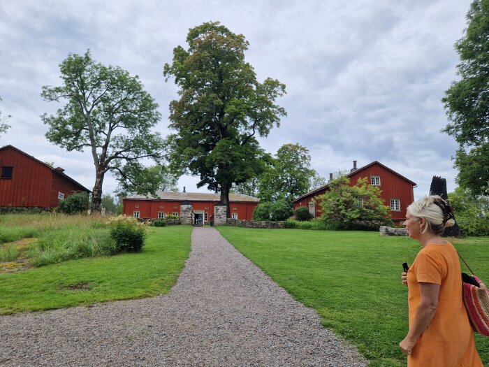 Kvinna på grusväg, röda träbyggnader, lummig trädgård, traditionell svensk gårdsmiljö, molnig himmel.