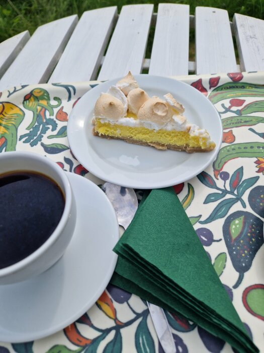 En bit paj med maräng på tallrik, kopp kaffe, utomhus på dekorerad duk, vita trädgårdsstolar i bakgrunden.