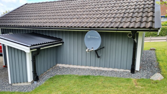 Carport i trä med satellitdisk, grå väggar, svart tak, grus, grönt gräs.