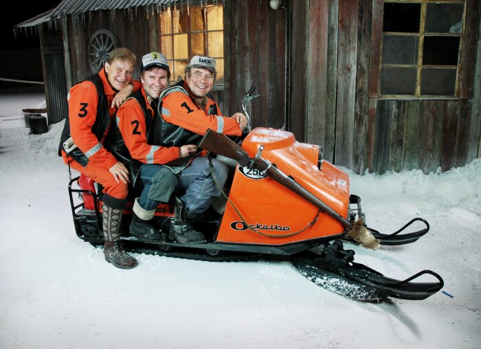 Tre leende män poserar på en orange snöskoter med nummerlappar framför en byggnad.