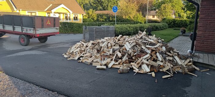 Stor hög med ved framför ett gult hus, trailer till vänster, asfalterad väg, cykelvägsskylt, grön häck.