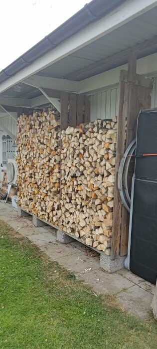 Vedstapel under tak vid husvägg, fläkt och svart tunna intill, gräsmatta i förgrunden.