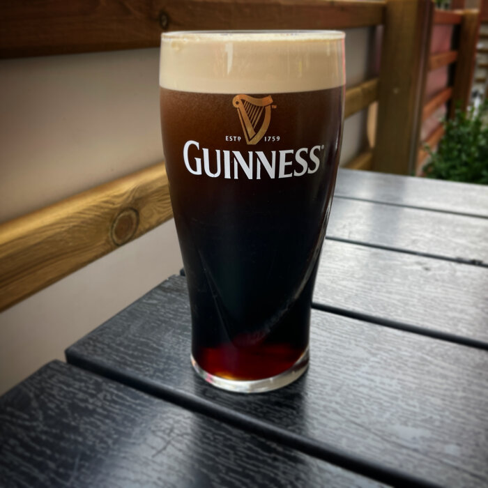 Ett fyllt Guinness ölglas på ett mörkt träbord utomhus. Skumtopp. Ljusdiffusion. Avslappnat.