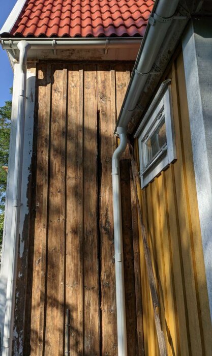 Trävägg, röd tegeltak, avloppsrör, fönster, gult hus, skuggor, soligt, utomhus, arkitektur, renoveringsbehov.