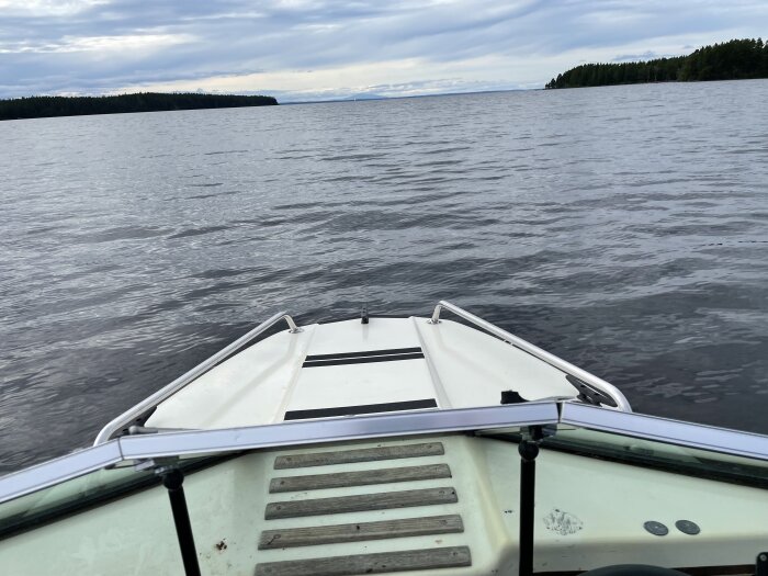 Framdel av båt glider fram på lugnt vatten, omgivet av skogklädda land och molnig himmel.