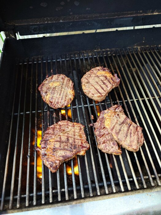 Fyra stekta köttbitar på en gasgrill med synliga lågor under.