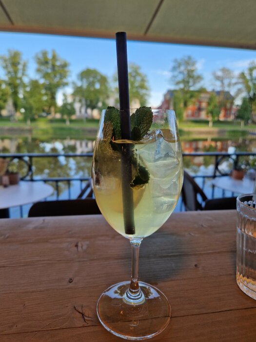 Ett glas med en drink, isbitar och myntablad, sugrör, uteservering med utsikt över park och byggnader, träbord.