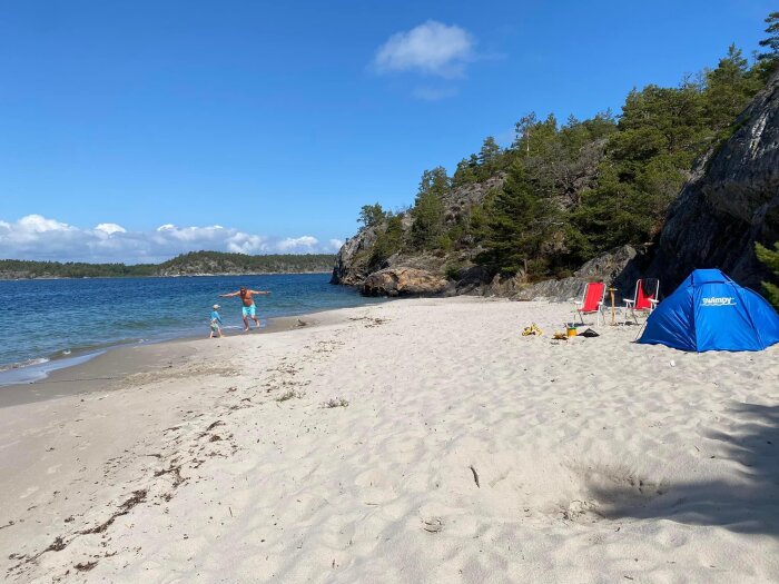 Stranddag, två personer promenerar, blå himmel, tält, stolar, skog, klippor.