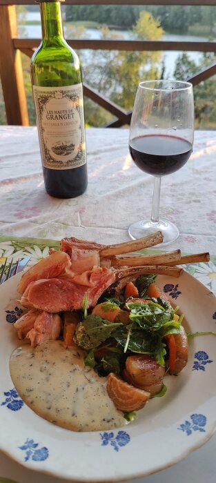 Middag utomhus med vin, lammkotletter, sallad, och sås på blommönstrad tallrik, naturutsikt i bakgrunden.