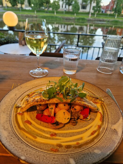 Måltid vid vattnet: fisken, grönsaker, aioli. Vinglas, upplagt på träbord, ute. Reflektion över flod, kvällsljus.
