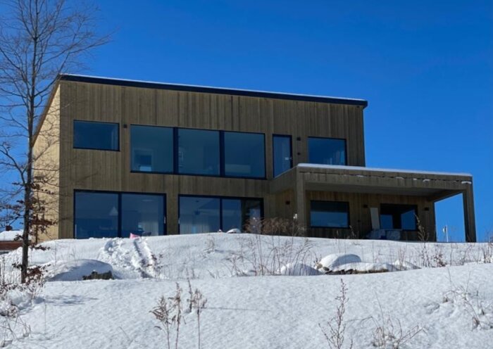 Modernt hus i snölandskap, stora fönster, klarblå himmel, träfasad, delvis begravd bil i snön.