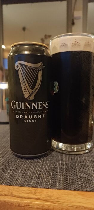 En Guinness ölburk bredvid ett halvfullt glas med mörkt stoutöl på en vävd bordstablett.