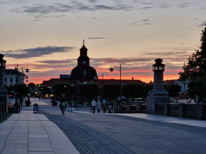 Stadsgata vid solnedgång, människor promenerar, cyklist, historisk byggnad, vacker himmel, färggranna moln.