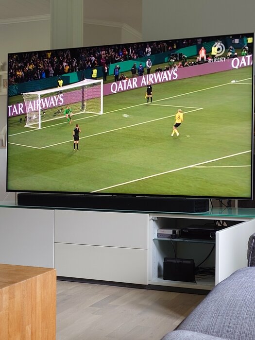 Fotbollsmatch på TV. Spelare förbereder sig för straff. Publik i bakgrunden. Inomhusmiljö.