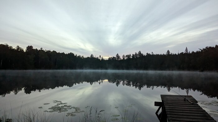 Morgondimma över stilla sjö, träbrygga, skogssilhuett, vattenspegling, strålande moln, gryningens ljus, lummig natur.