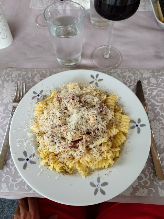 En tallrik pasta med riven ost, svartpeppar, kniv, gaffel, glas vatten och rödvin, dukat bord.