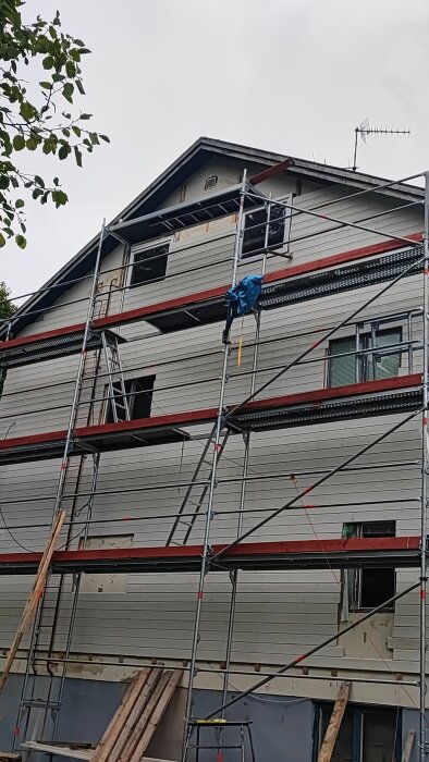 Ett hus med ställningar runtom, renoveringsarbete pågår, grå molnig himmel. Trädgren i förgrunden.