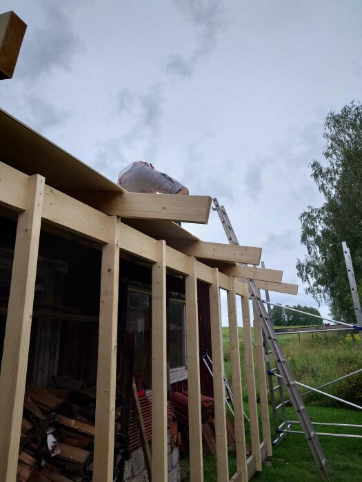 Person jobbar på ett tak till en träkonstruktion, molnig himmel, stege i förgrunden, grönt gräs baktill.