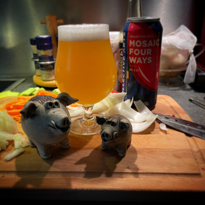 Ölglas med skum, grishantverk, burk, skärbräda, kniv, grönsaker, köksmiljö.