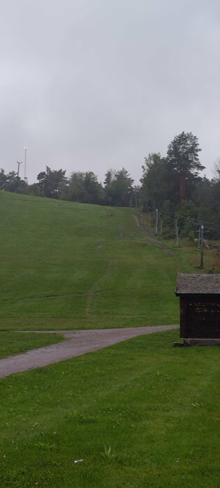 En gråmulen dag, grönt gräs, en liten stuga, gångvägar, lyktstolpar, träd, en kulle, inga personer synliga.