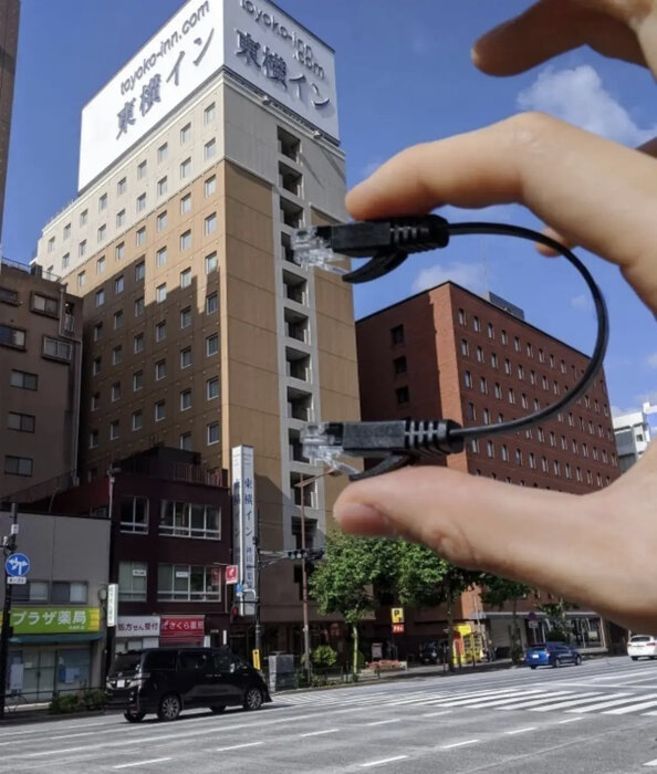 En hand håller en nätverkskabel, skapar optisk illusion med en byggnad i bakgrunden.