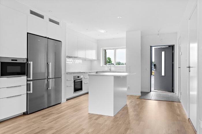 Modernt kök med vit inredning, rostfritt kylskåp, inbyggd ugn och trägolv. Ljust och minimalistiskt.