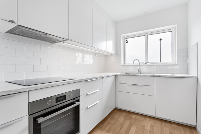 Modernt, minimalistiskt kök i vitt med ugn, spishäll, diskho och fönster.