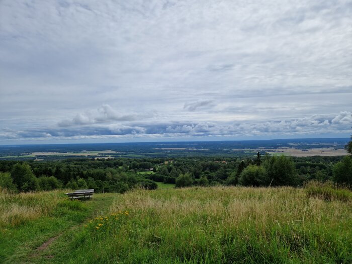 Panoramavy över landskap med molnig himmel, träd, åkrar och en ensam bänk i förgrunden.