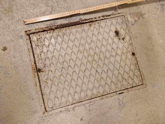 Metallucka i golv, skarpt ljus, måttstock ovanpå, rostiga fläckar, betongyta runt omkring, industriell eller källarmiljö.