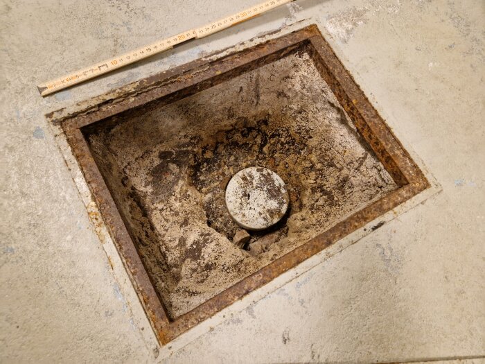 Öppen kvadratisk brunn, rostiga kanter, måttstock ovanpå, betonggolv, grus och smuts inuti.