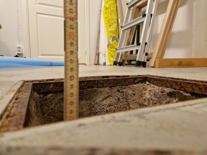 Tumstock i öppet hål i golv, inomhus byggarbetsplats, vit dörr i bakgrunden, stege till höger.