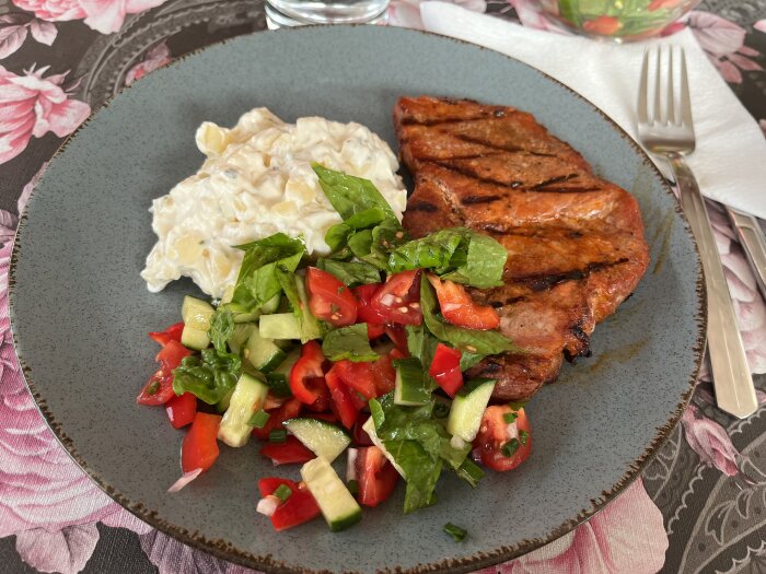 Grillad köttbit, färsk sallad med grönsaker, potatissallad, matplats med blommig duk.