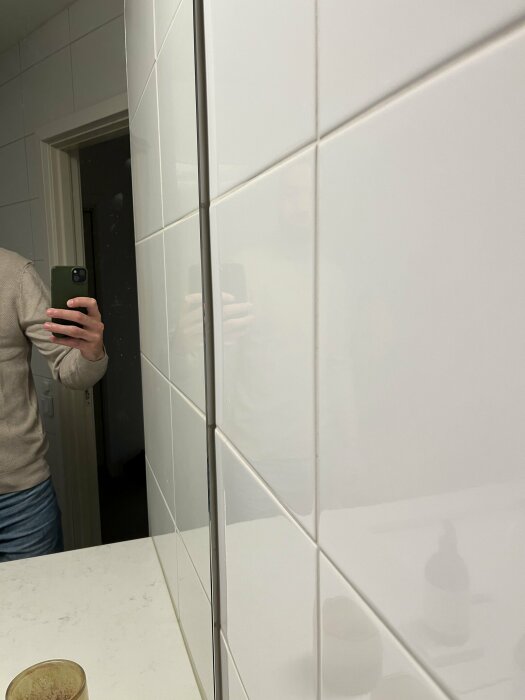 En person tar en selfie i en spegel i ett badrum med vita kakelväggar.