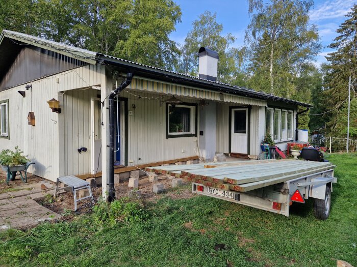 Ett hus med veranda under konstruktion, skottkärra, släpvagn, och trädgårdsredskap på gräsmattan.