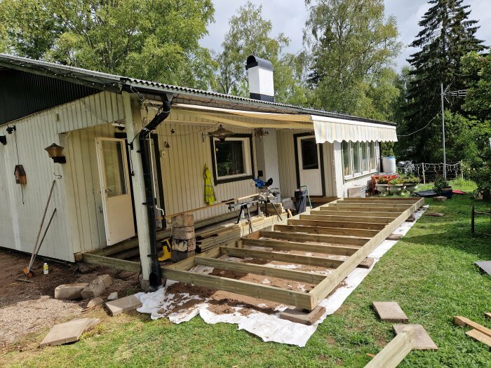 Pågående bygge av träterrass vid hus, verktyg och byggmaterial synliga, grönska i bakgrunden, dagtid.