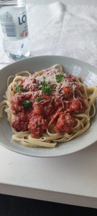 En tallrik spaghetti med köttfärssås och riven ost, en vattenflaska i bakgrunden, på ett vitt bord.