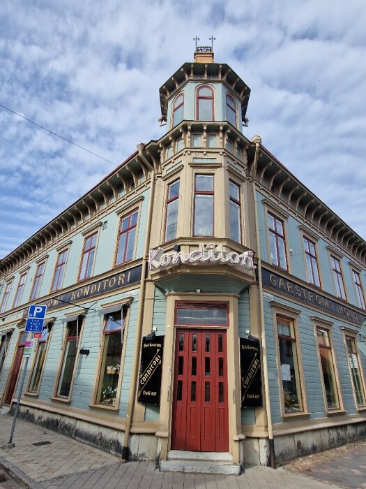 Hörnbyggnad med konditori, tegeltak, torn, röda dörrar, fönsterluckor, och skyltar på gatunivå, mot molnig himmel.