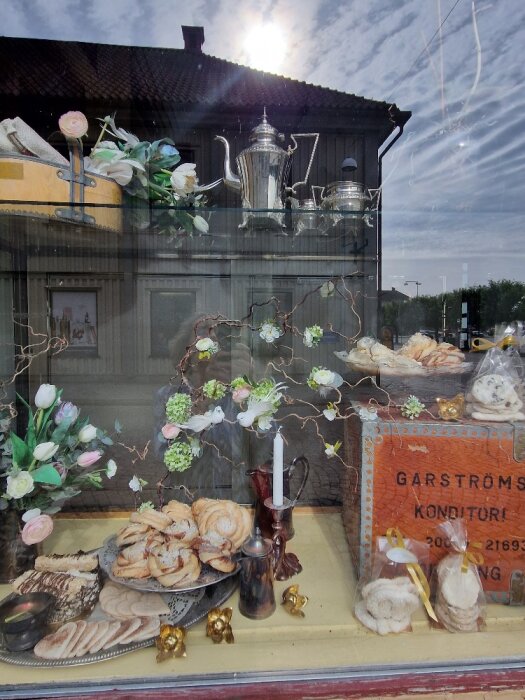 Ett konditoriskyltfönster med blommor, bakverk, ljus och kaffekannor. Reflektion av byggnad och himmel syns.